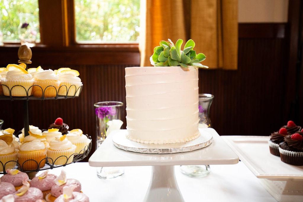 A simple white wedding cake with a succulent on top sits on a table of treats.