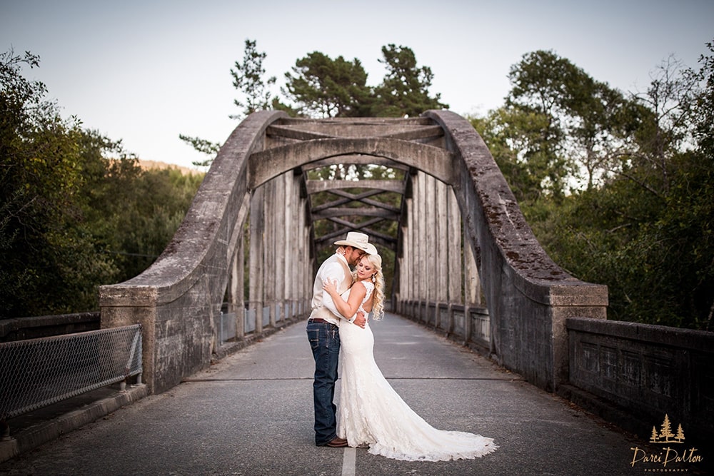 A bride and groom embrace on a concrete bridge with green trees in the background.