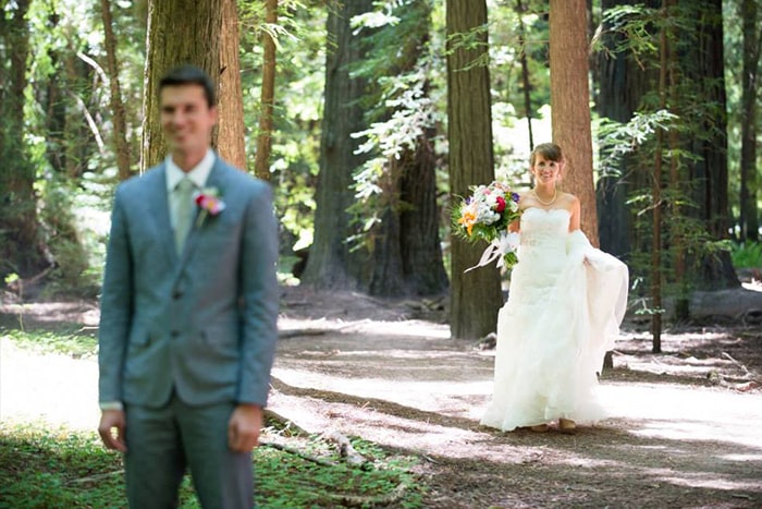 A groom stands with his back to his bride as she slowly approaches him for their first look. Tall redwood trees surround them.